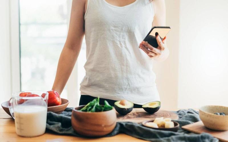 Cropped shot of a young woman checking recipe on smartphone while preparing ingredients to make a healthy smoothie with fruits, vegetables and vegan milk.